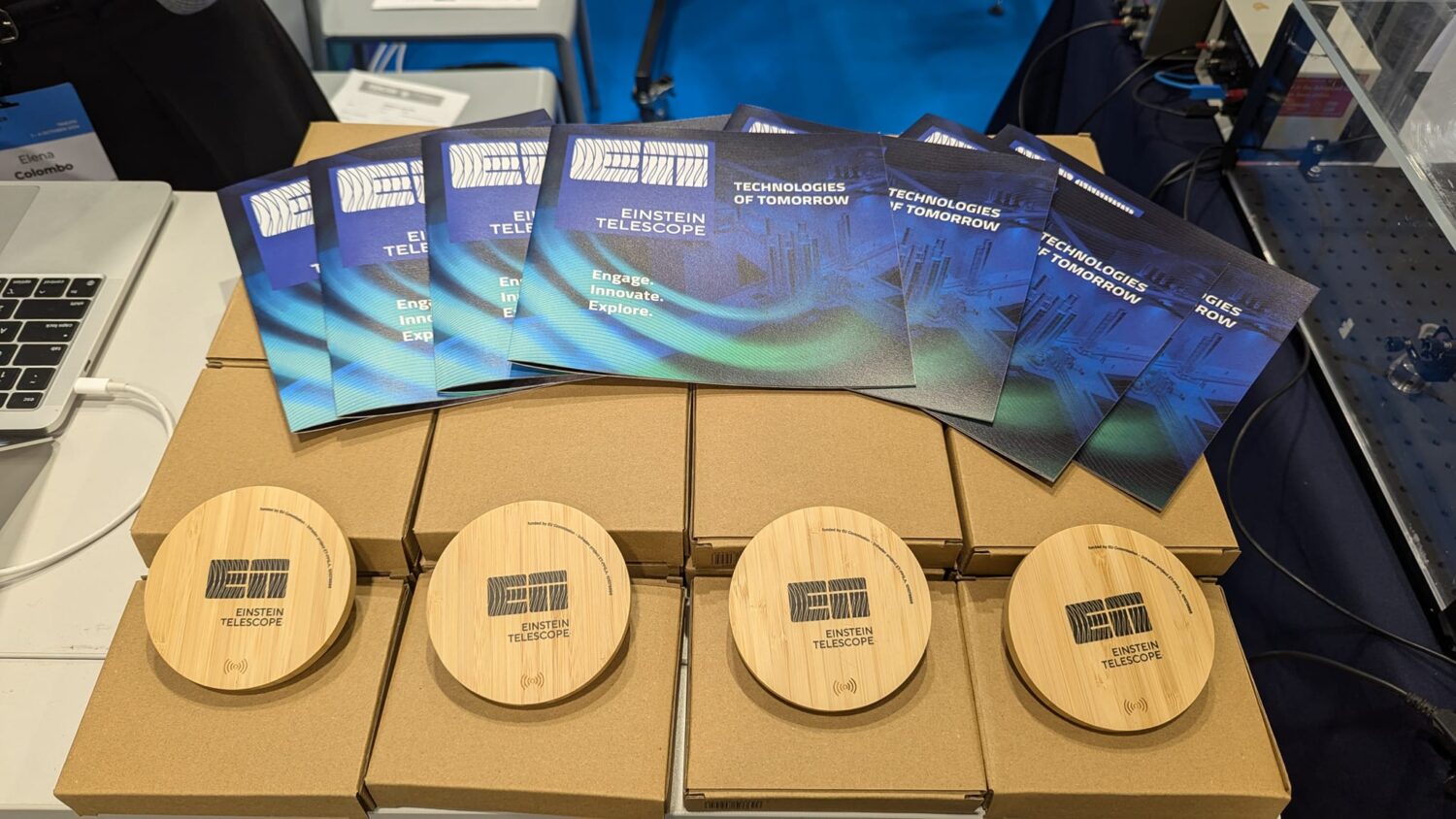 The photo shows brochures and wireless chargers with the Einstein Telescope logo.
