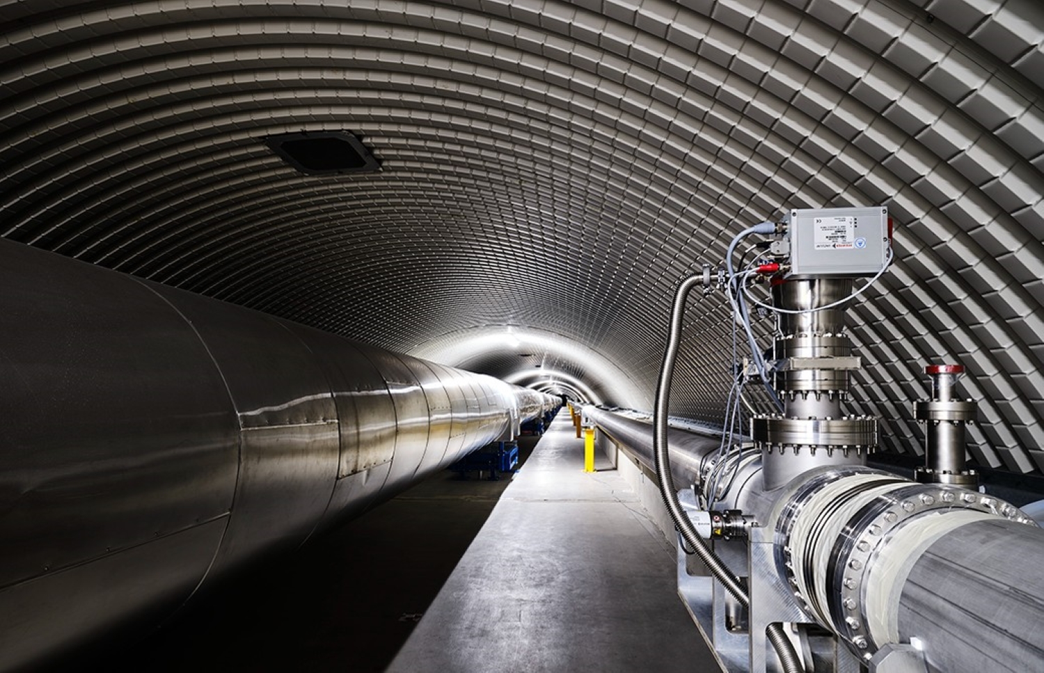 A view inside a large, curved tunnel housing sections of a gravitational wave detector. The tunnel contains long, metallic vacuum tubes supported by blue mounts, with technical instrumentation and cables visible in the foreground. The space is brightly lit with a repeating overhead light pattern. Credits: Enrico Sacchetti