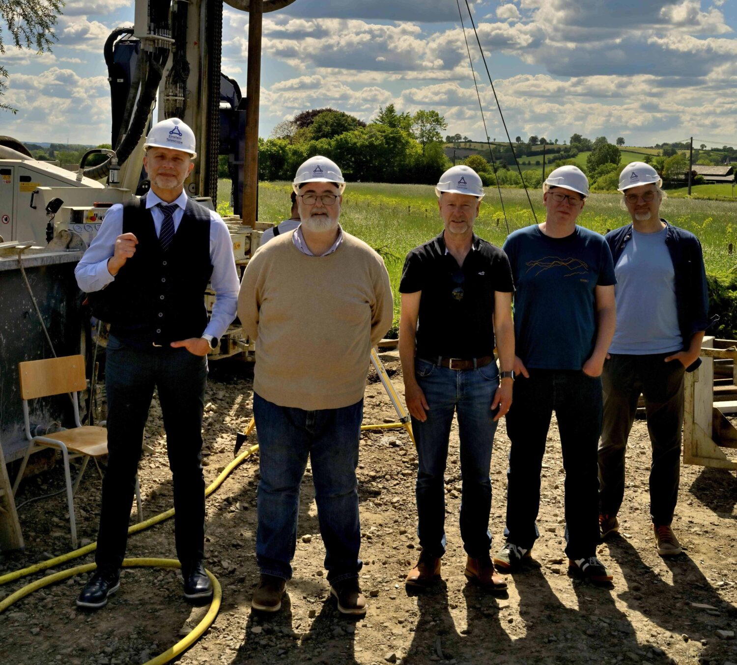 Five men with white helmets standing in front of a drilling machine and a green landscape with a sky and clouds above it.