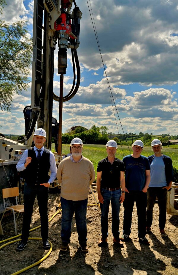 Five men with white helmets standing in front of a drilling machine and a green landscape with a sky and clouds above it.