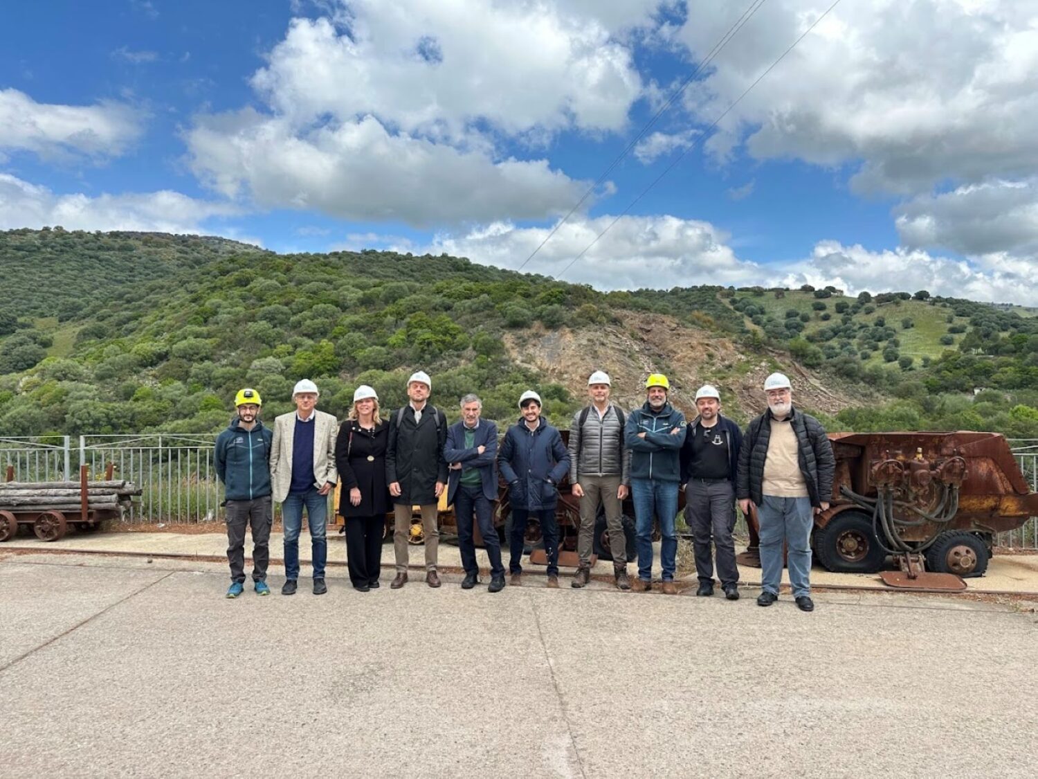 Ten people, most of them wearing helmets, in front of a green hill with a blue sky with white coulds above it.