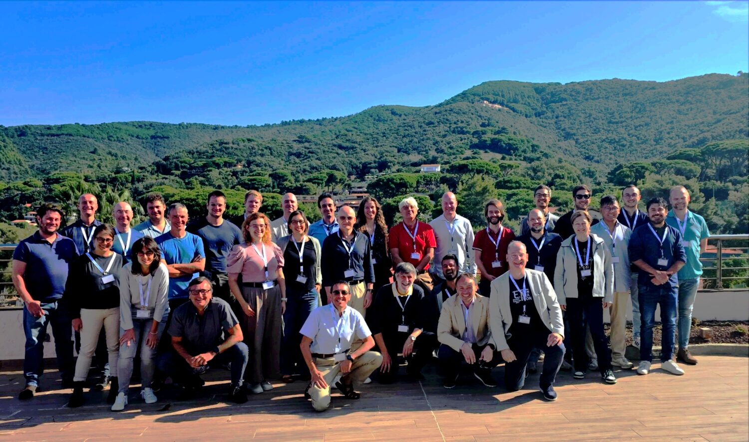 Group photo of the workshop participants on an outdoor terrace: about thirty people are standing and sitting in several rows, many wearing name badges on lanyards. In the background are green hills under a bright sky and a distant line of sea; a sunny day with a panoramic view.