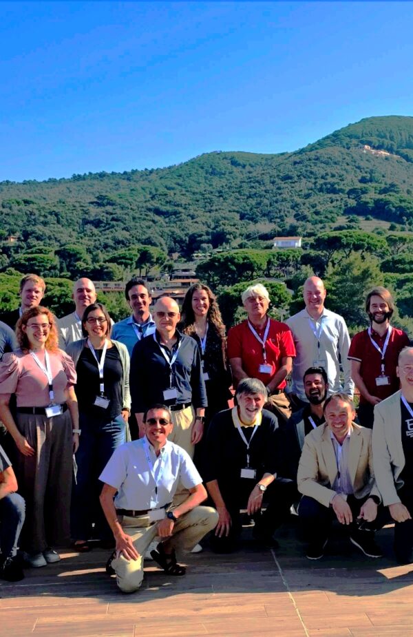 Group photo of the workshop participants on an outdoor terrace: about thirty people are standing and sitting in several rows, many wearing name badges on lanyards. In the background are green hills under a bright sky and a distant line of sea; a sunny day with a panoramic view.
