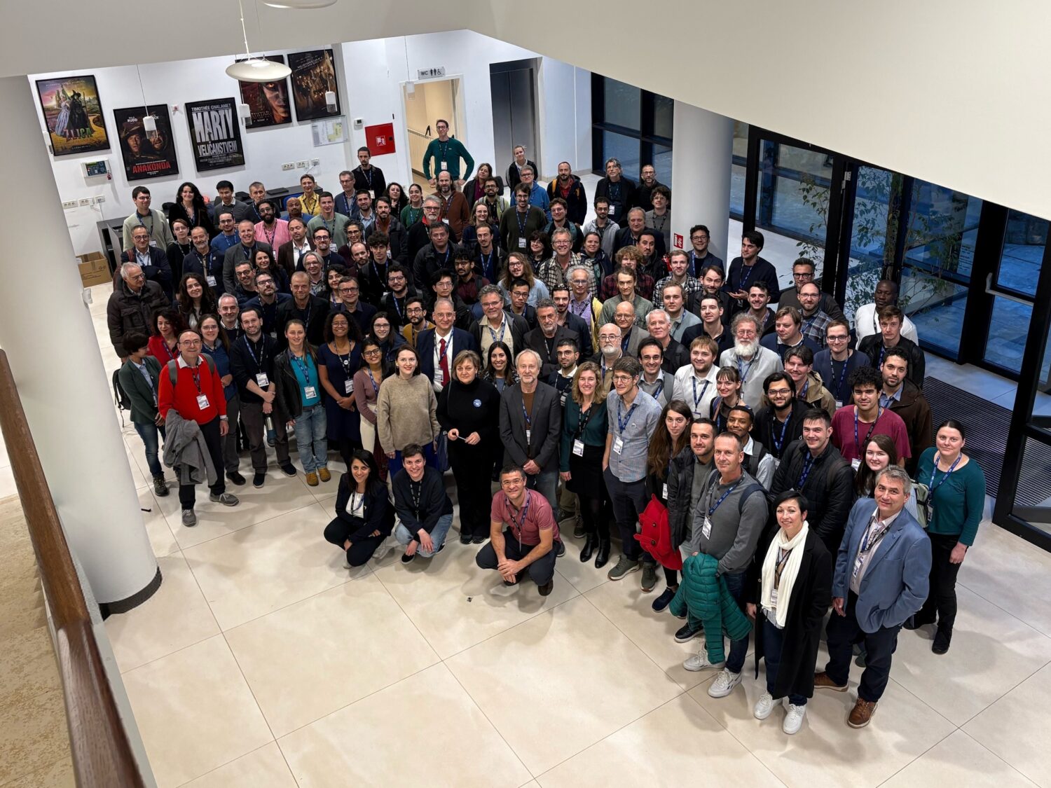A large group of Einstein Telescope Annual Meeting participants gathered indoors for a group photo, standing closely together and smiling at the camera. The photo is taken from an elevated viewpoint in a bright lobby with posters on the wall and large windows in the back.