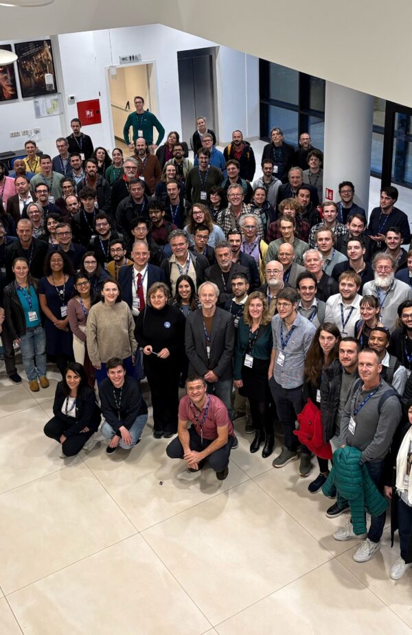A large group of Einstein Telescope Annual Meeting participants gathered indoors for a group photo, standing closely together and smiling at the camera. The photo is taken from an elevated viewpoint in a bright lobby with posters on the wall and large windows in the back.