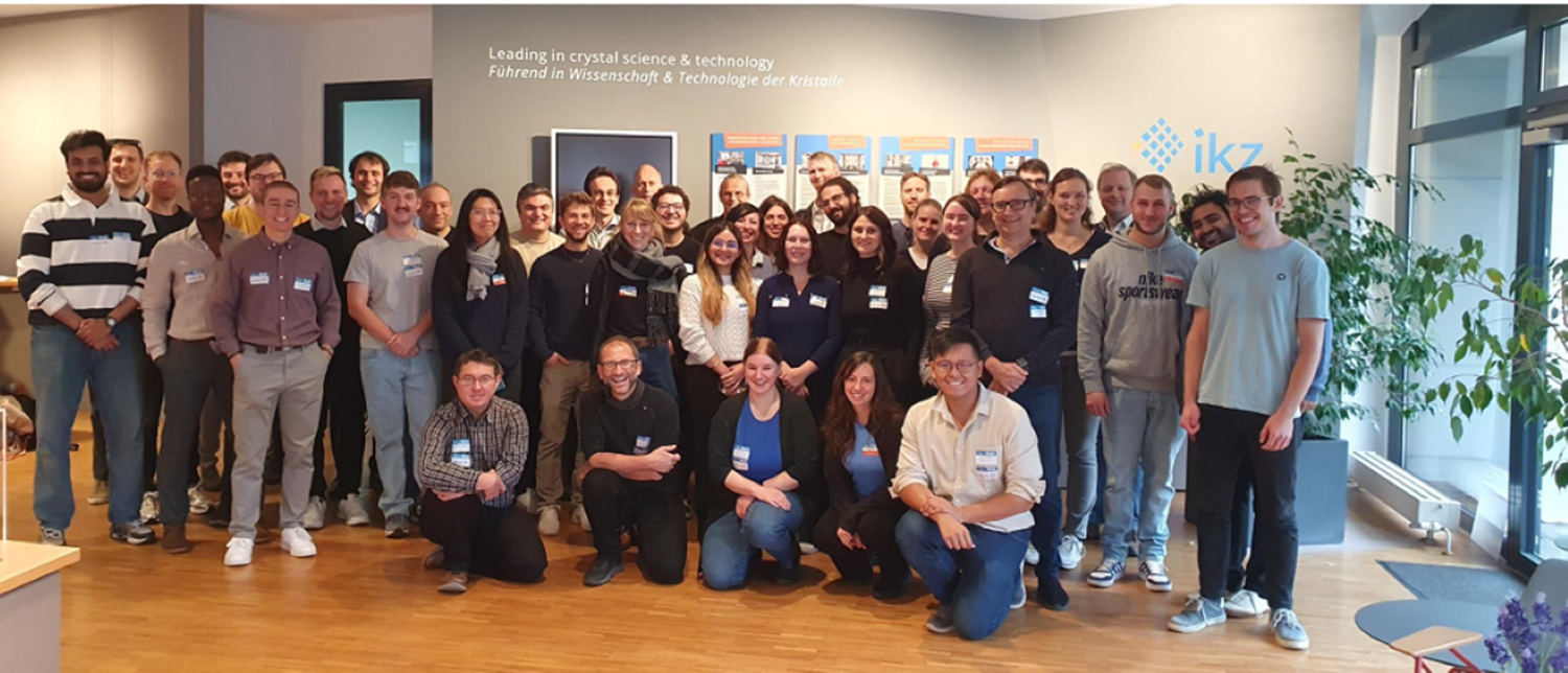 Large group photo taken indoors at a research institute: around 35–40 conference participants of mixed genders stand and kneel in several rows, all wearing name badges and smiling at the camera. Behind them are posters on the wall and the “ikz” logo; the room has wooden floors, bright lighting, and a large potted plant by the windows on the right.
