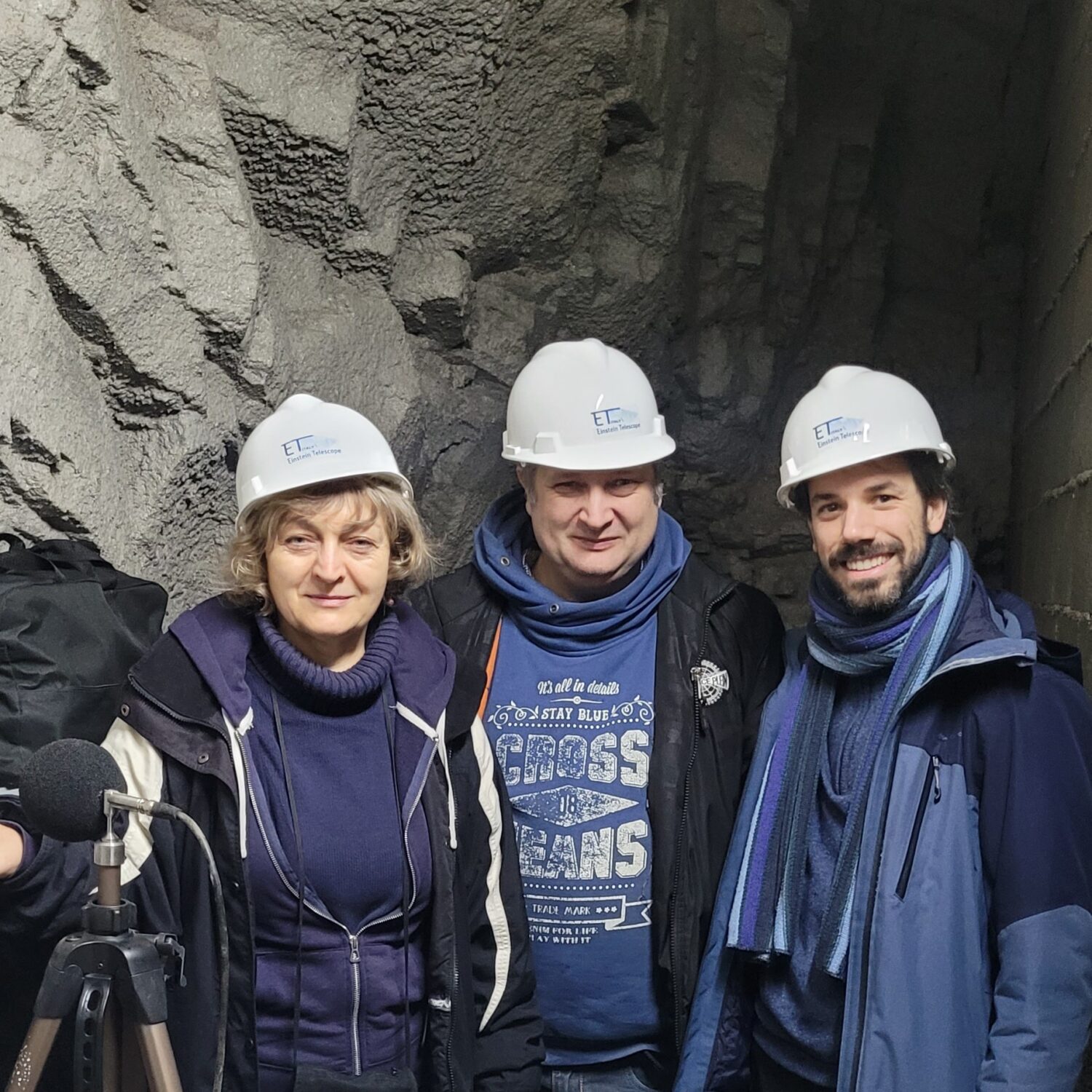 Three people wearing white hard hats pose inside a rocky underground mine tunnel; a microphone setup is visible in the foreground.