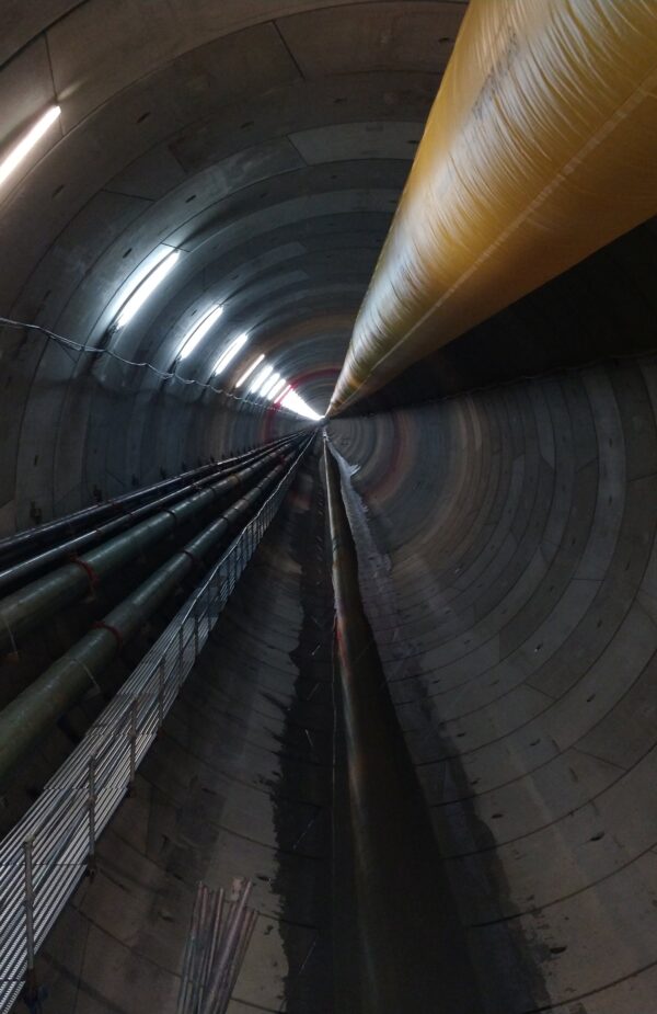A long, cylindrical underground tunnel lined with lights, with large pipes and cables running along the walls and ceiling toward a distant vanishing point.