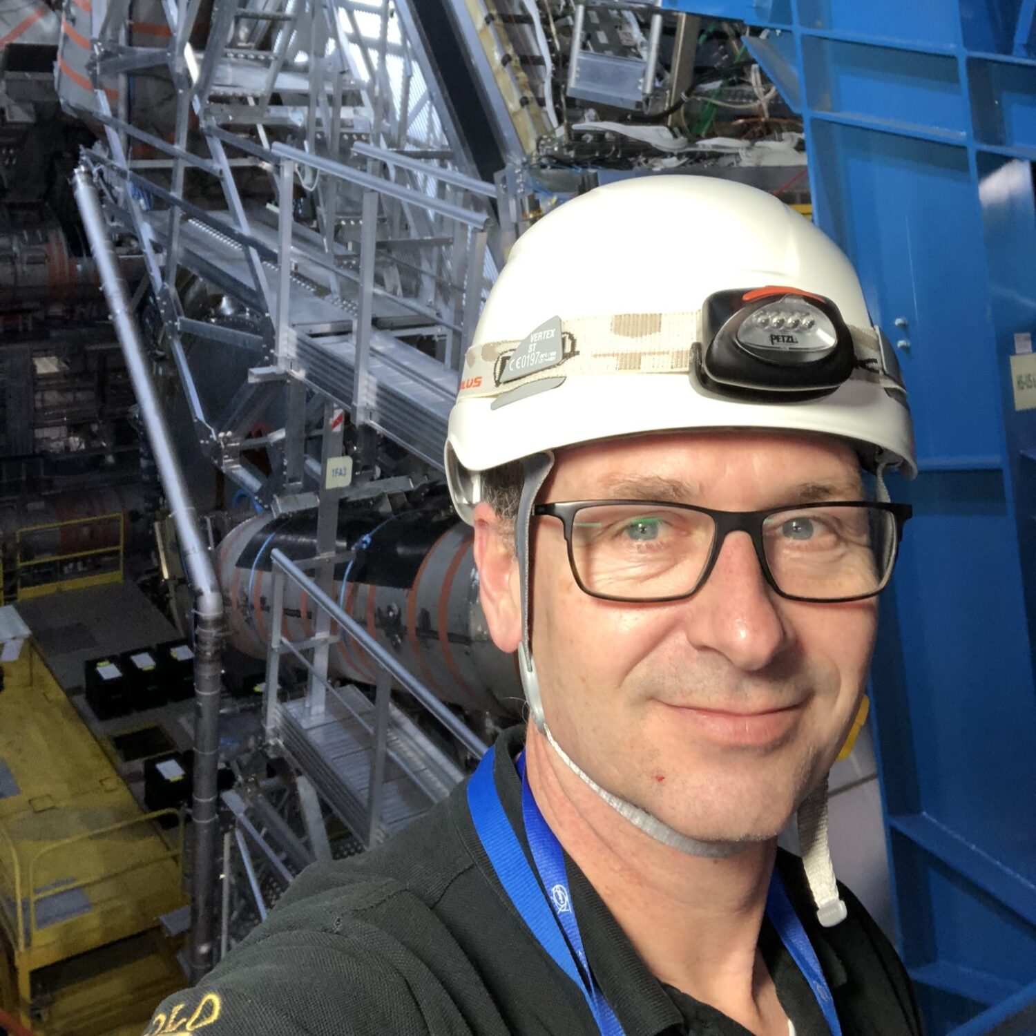 A person wearing glasses and a white hard hat with a headlamp takes a selfie inside an industrial facility, with metal platforms, pipes, and large machinery in the background.
