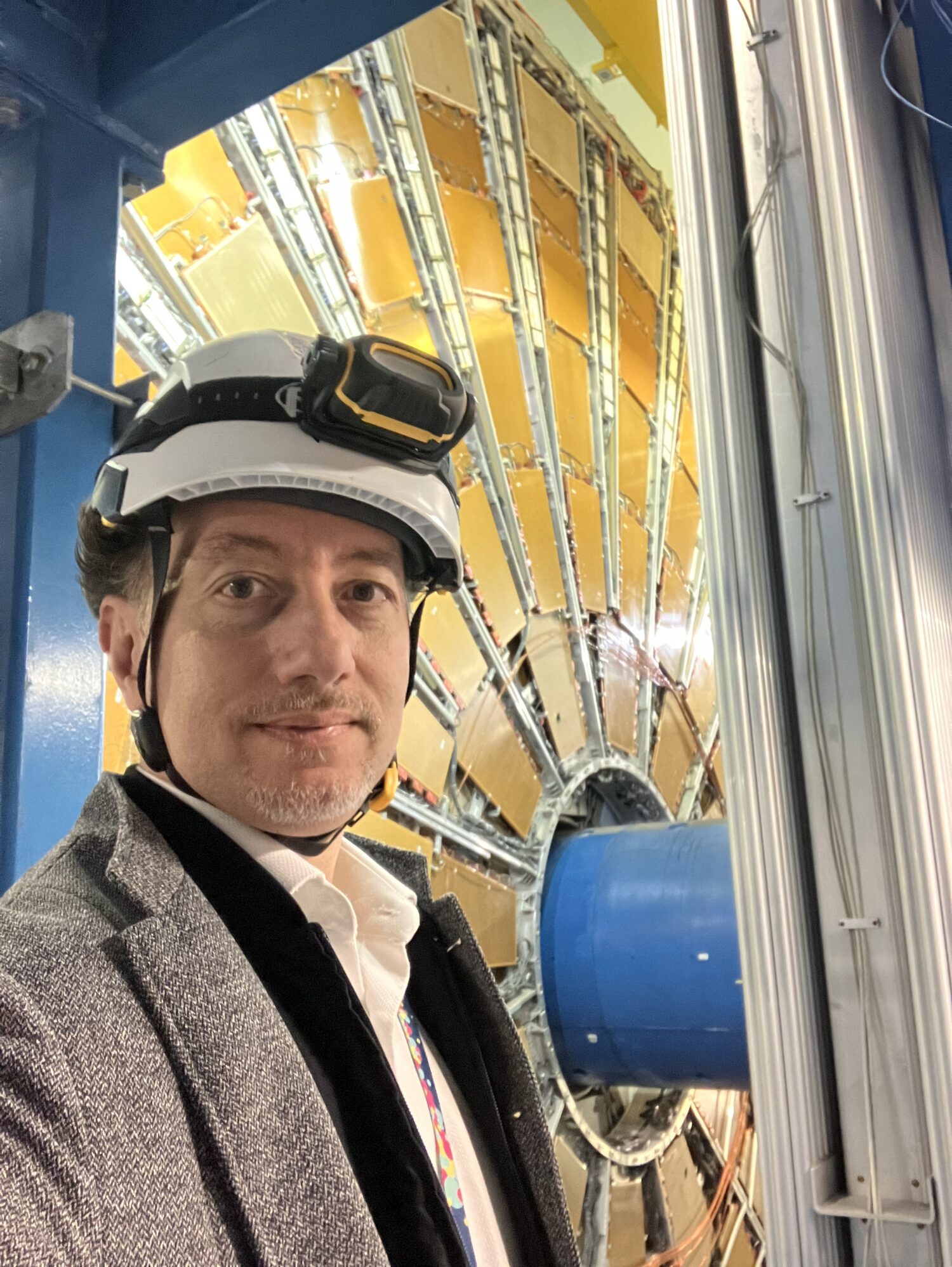 A person wearing a white hard hat with protective goggles takes a selfie inside an industrial research facility, with a large circular detector structure and blue machinery behind them.