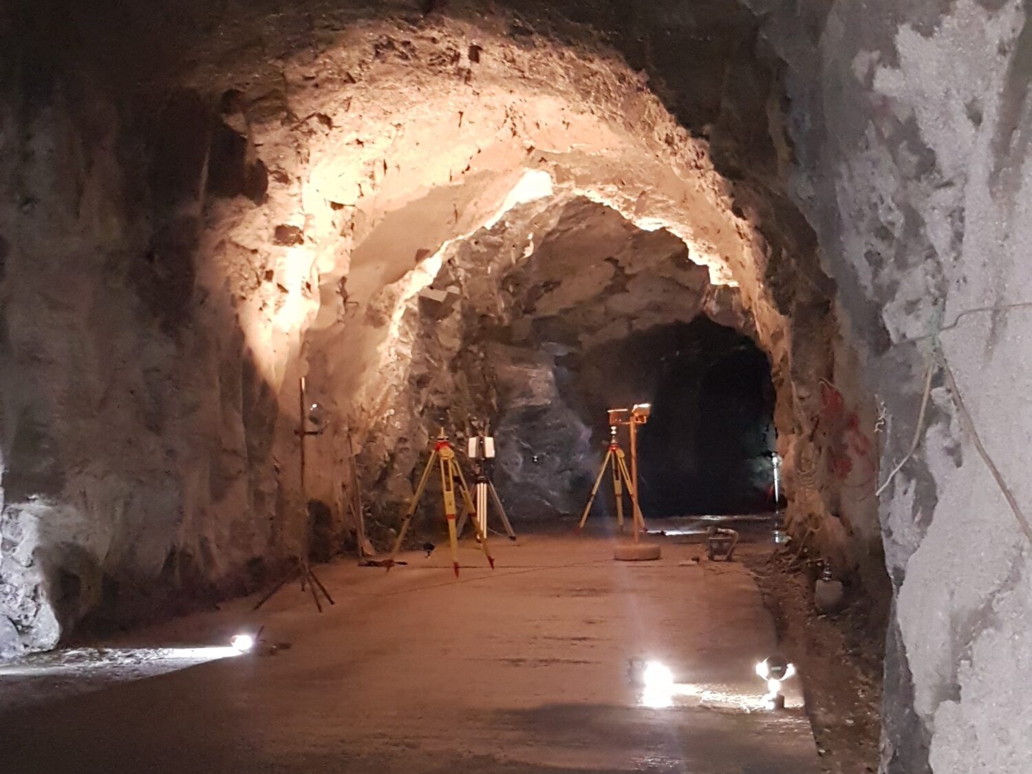 A wide view of a rocky underground tunnel lit by floor lamps, with several tripods and measurement instruments set up along the passage.