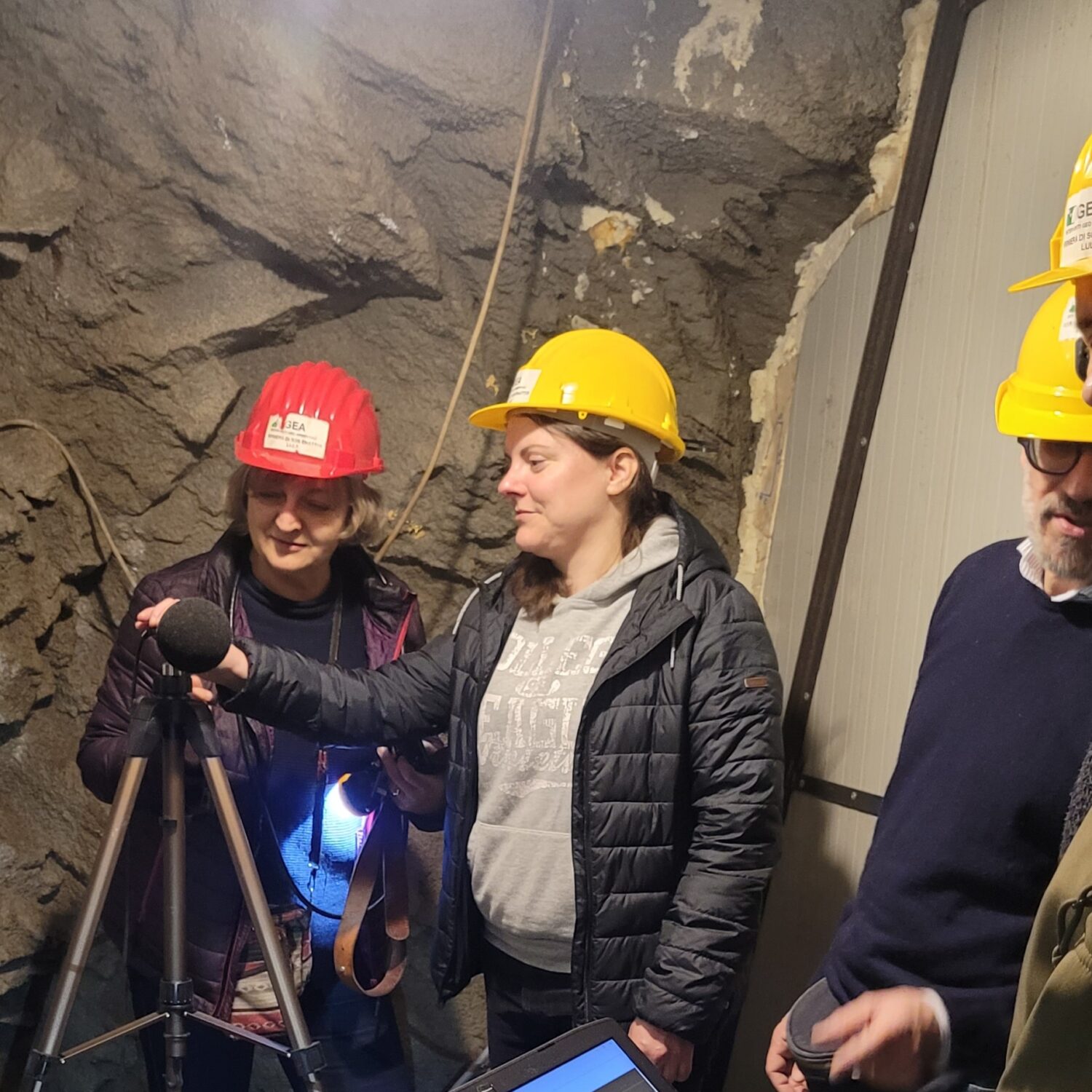 Two people wearing hard hats stand in a rocky underground mine tunnel, adjusting a microphone on a tripod; one holds a headlamp that illuminates the scene.